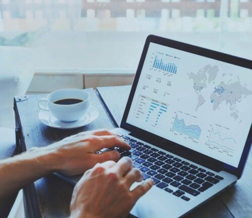 Hands typing on laptop displaying financial dashboard with charts, graphs, and world map