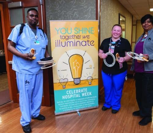 Three healthcare workers celebrating Hospital Week with food and promotional banner