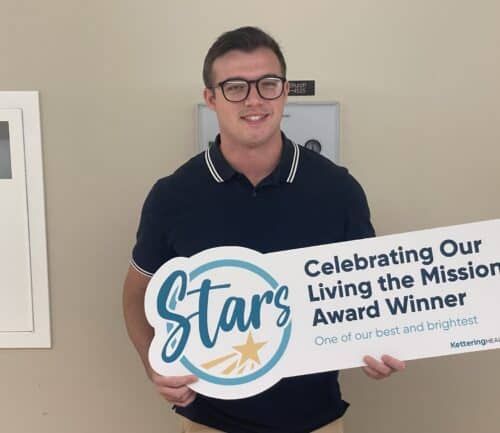 Male healthcare employee holding "Stars Celebrating Our Living the Mission Award Winner" sign