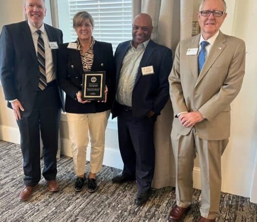 Four business professionals pose together, woman holding award plaque at formal ceremony