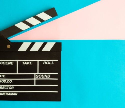 Black film clapperboard with white diagonal stripes on bright blue and pink background.