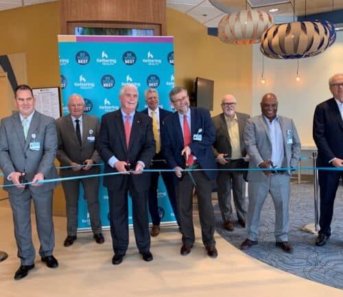 Eight men in business suits holding scissors for ribbon cutting ceremony at healthcare facility opening