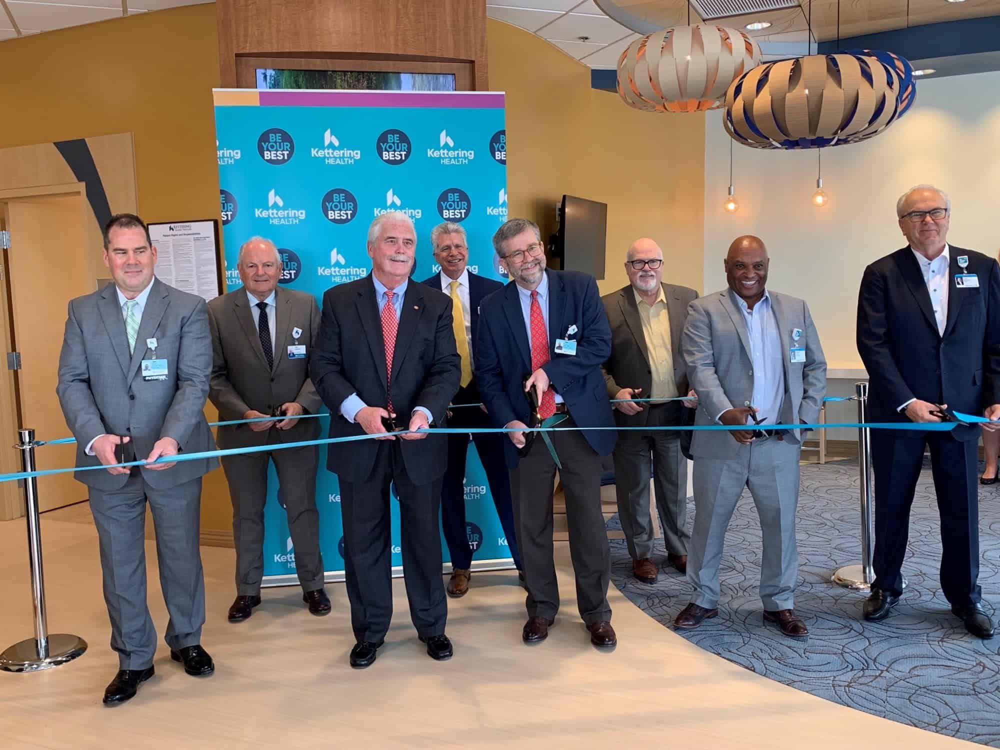 Eight men in business suits holding scissors for ribbon cutting ceremony at healthcare facility opening
