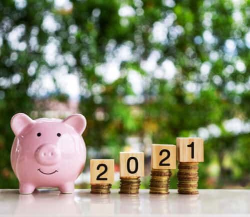 Pink piggy bank beside stacked coins with 2021 wooden blocks on white surface