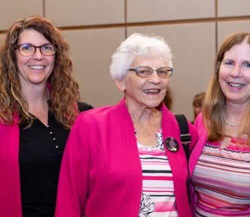 Three women in pink blazers smiling together at healthcare event