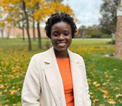 Professional headshot of Raina Simon standing outdoors in autumn wearing orange top and beige coat