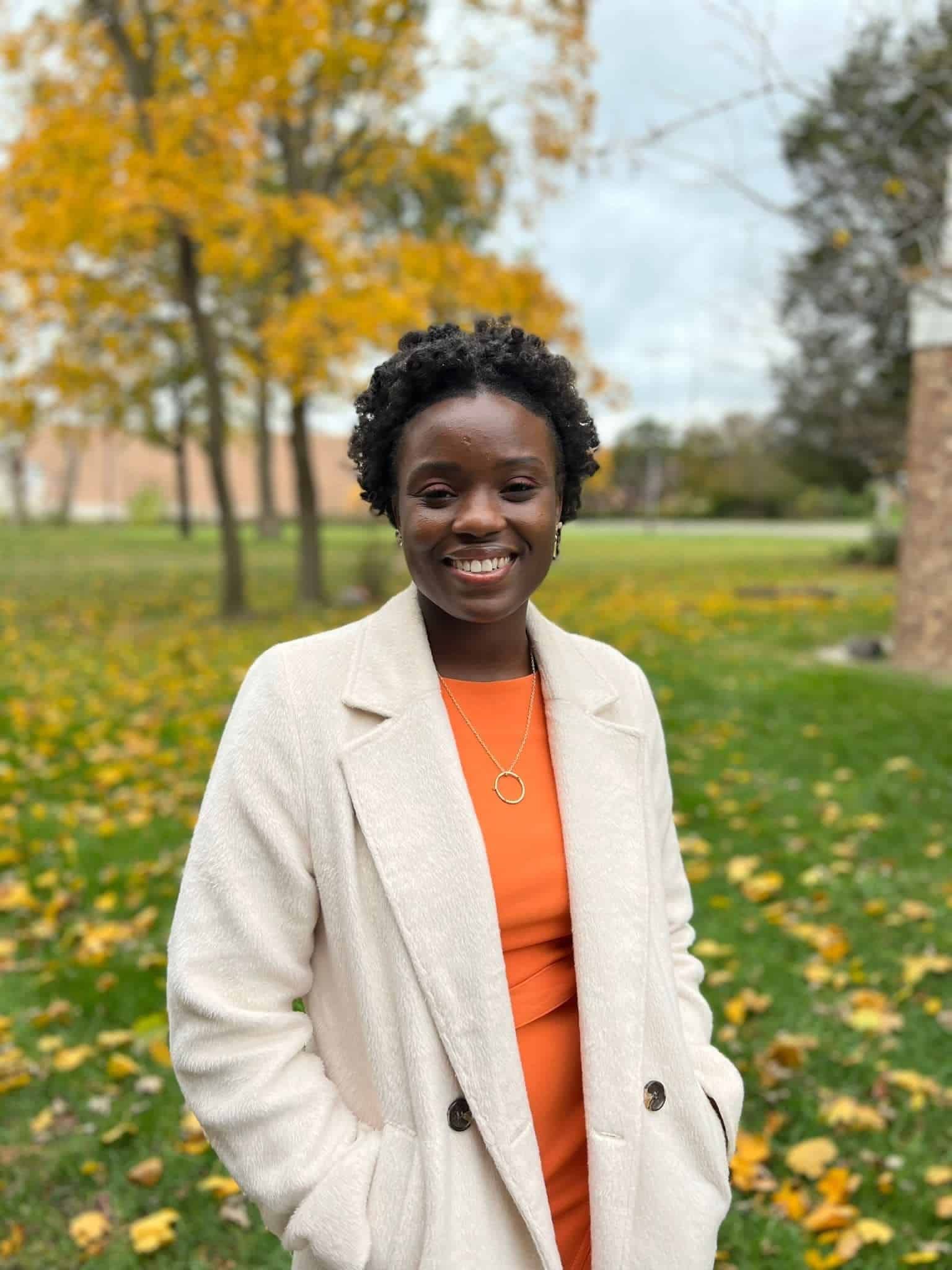 Professional headshot of Raina Simon standing outdoors in autumn wearing orange top and beige coat