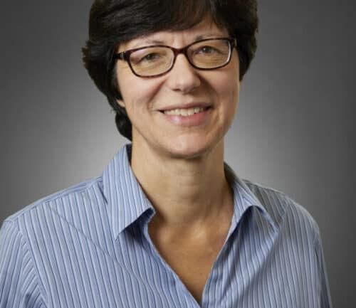 Professional headshot of Randi Quale smiling, wearing glasses and blue striped shirt