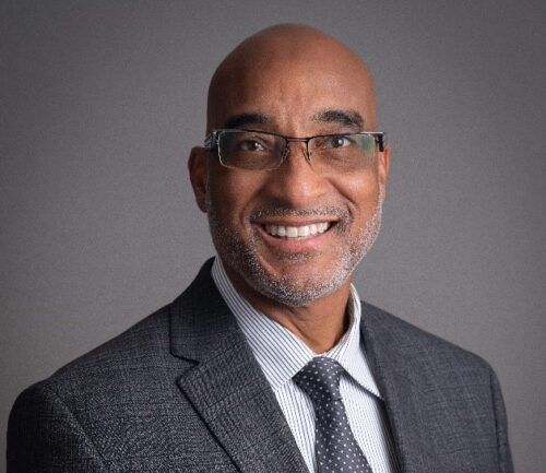 Professional headshot of Ron Mainess in suit and glasses, smiling at camera