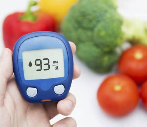 Hand holding blood glucose meter showing 93 mg/dL with fresh vegetables in background