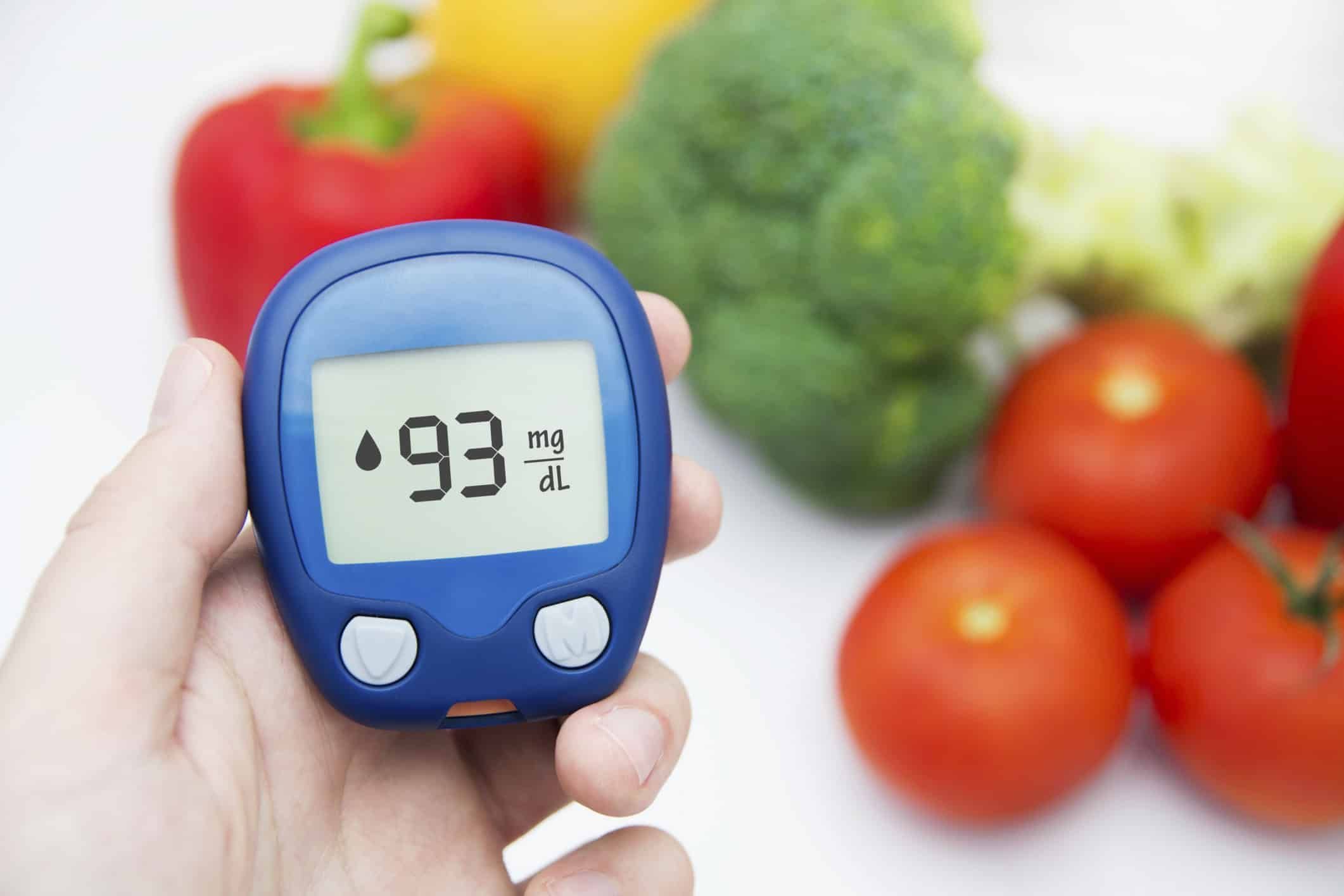 Hand holding blood glucose meter showing 93 mg/dL with fresh vegetables in background
