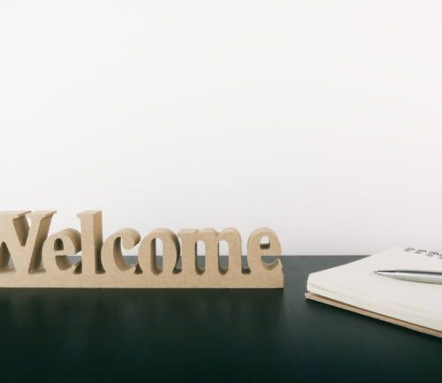 Wooden "Welcome" sign on dark desk beside open notebook and pen