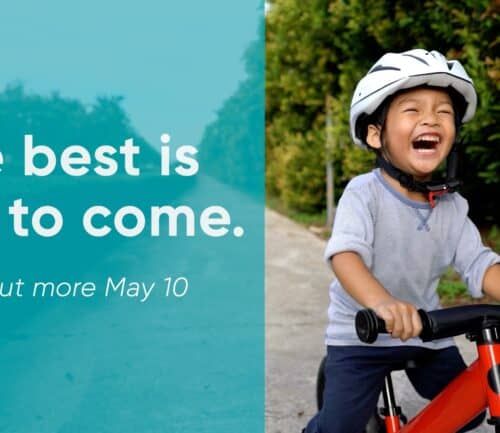 Young child laughing while riding red bike wearing white helmet with "The best is yet to come" text