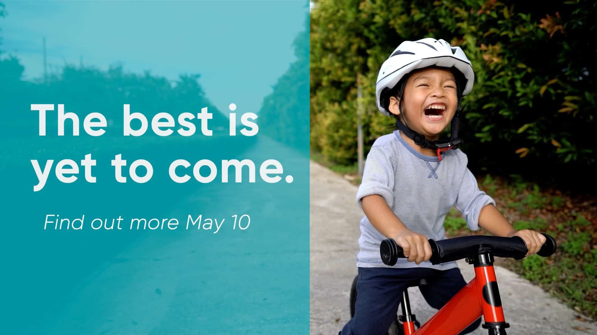 Young child laughing while riding red bike wearing white helmet with "The best is yet to come" text