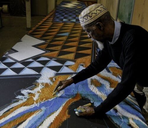 Elderly man in traditional cap painting colorful geometric artwork on large canvas