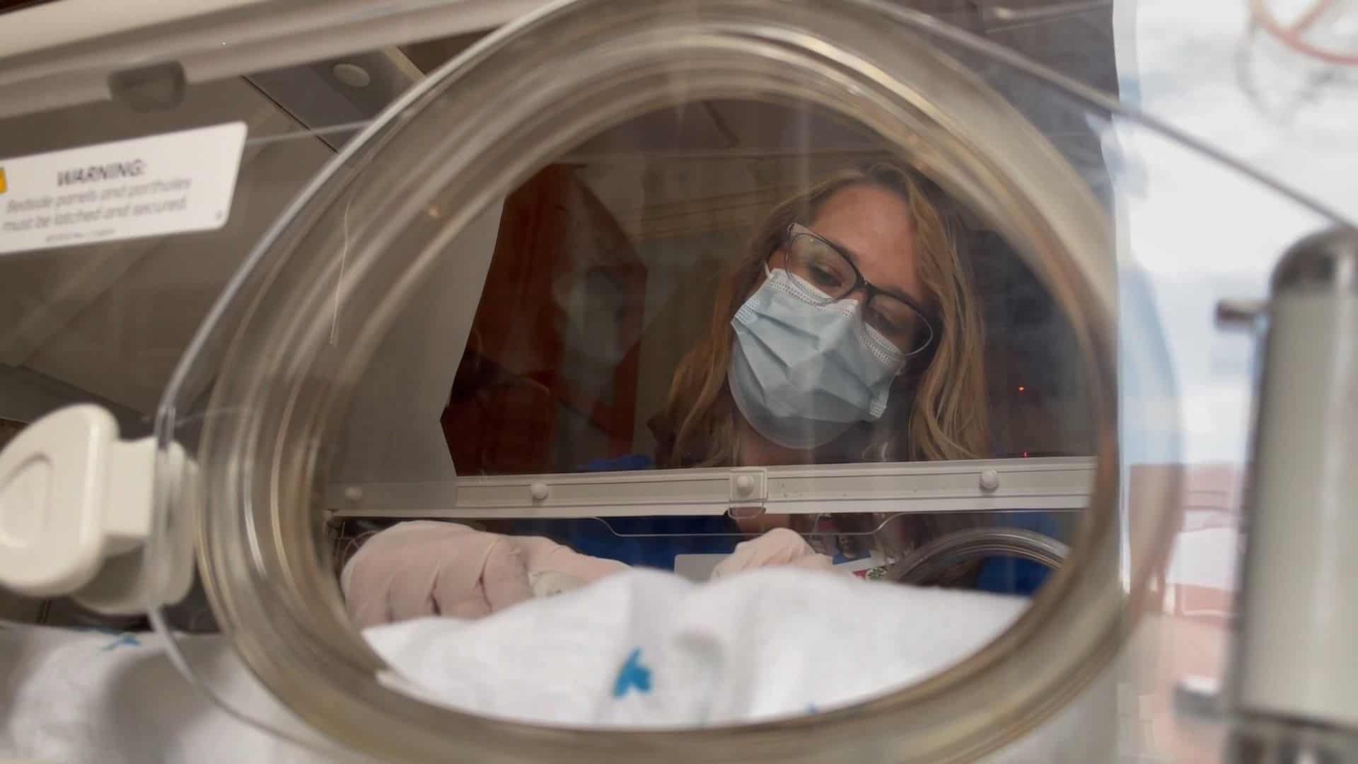 Healthcare worker in protective mask examining newborn baby in hospital incubator