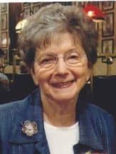 Elderly woman with glasses and gray hair smiling warmly in blue cardigan