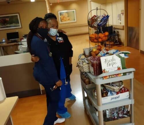 Two healthcare workers in scrubs embracing beside mobile cart with snacks and supplies