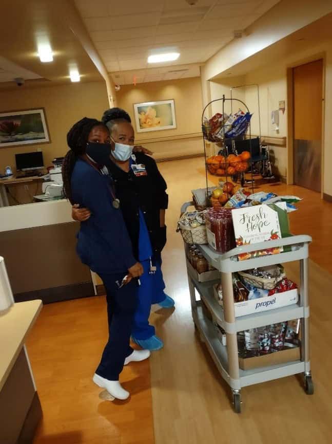 Two healthcare workers in scrubs embracing beside mobile cart with snacks and supplies