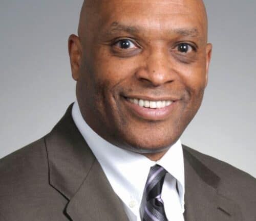 Professional headshot of George Lewis in gray suit with striped tie smiling