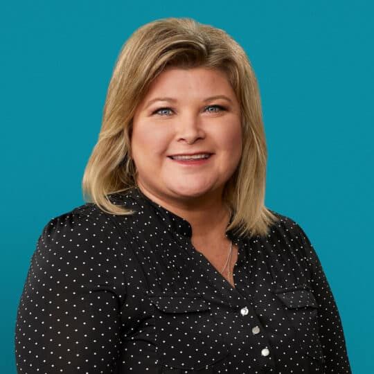 Professional headshot of Joann Heuker, APRN-CNP, smiling in polka dot blouse against teal background.