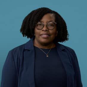 Professional headshot of Karlene Williamson wearing glasses and navy blazer against blue background