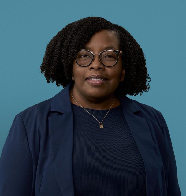 Professional headshot of Karlene Williamson wearing glasses and navy blazer against blue background