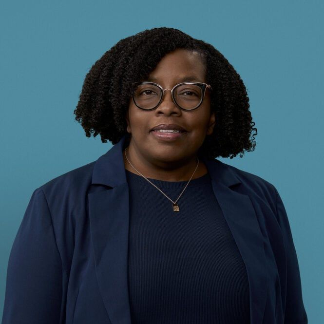 Professional headshot of Karlene Williamson wearing glasses and navy blazer against blue background