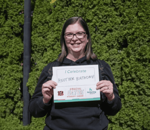 Woman holding sign reading "I Celebrate Another Birthday!" outdoors against green hedge