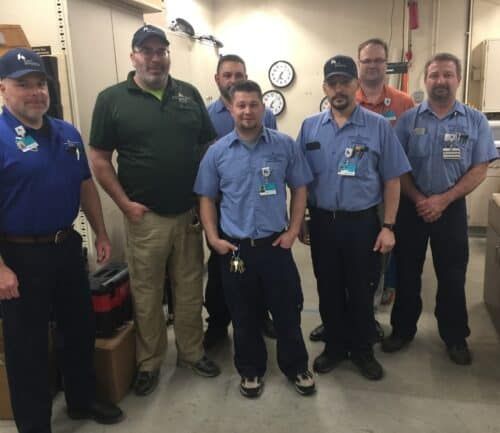 Seven Kettering Health plant engineering staff members posing together in maintenance workshop facility.