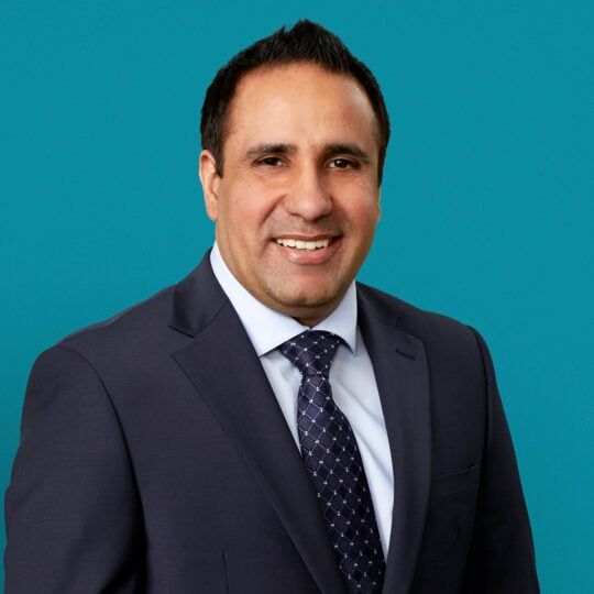 Professional headshot of Dr. Sukhchain Singh in dark suit against teal background