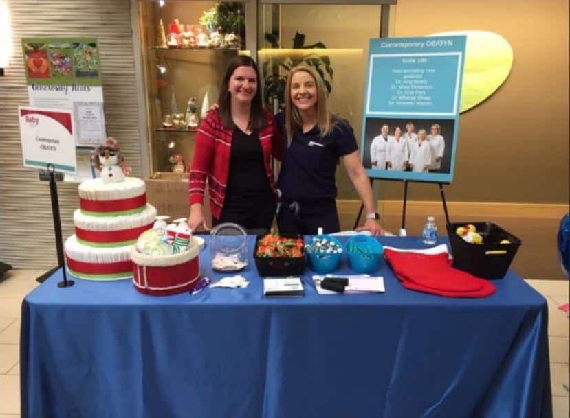Two healthcare staff members standing behind informational table with cake and promotional materials