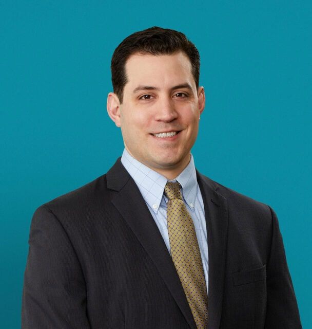 Professional headshot of Dr. Zoltan Krudy in dark suit against teal background