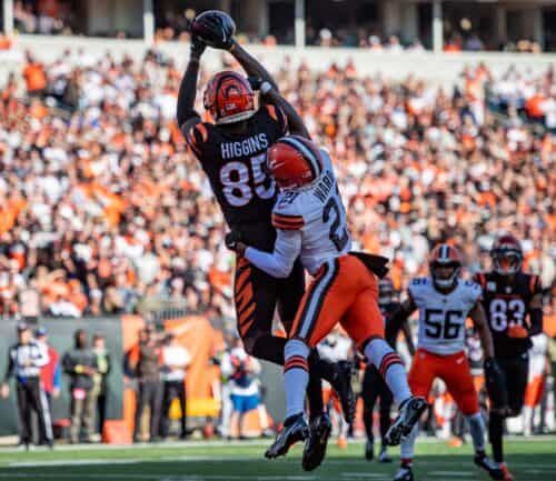 Football player in black uniform catches ball while defender in orange attempts tackle