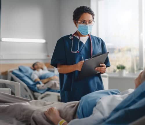 Nurse in blue scrubs and mask checks on elderly patient in hospital bed