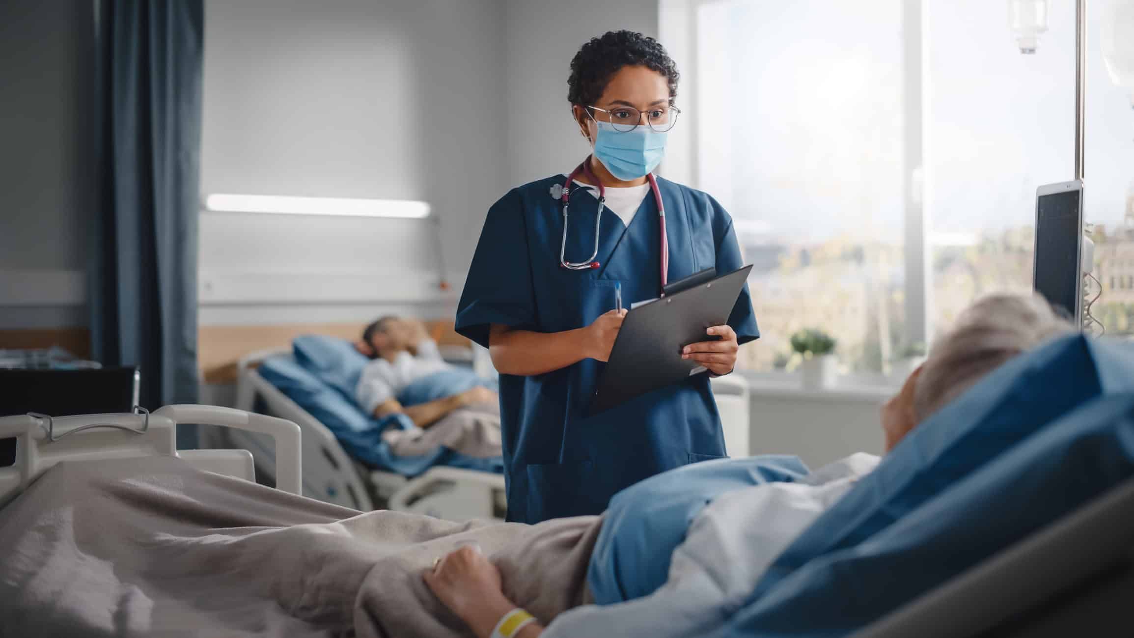 Nurse in blue scrubs and mask checks on elderly patient in hospital bed