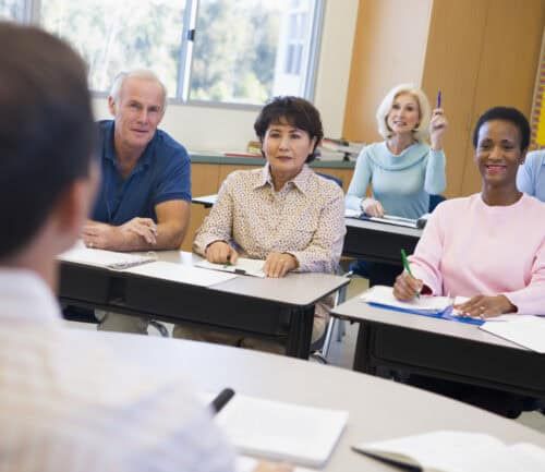 Mature adults sitting at desks in classroom, woman raising hand to participate