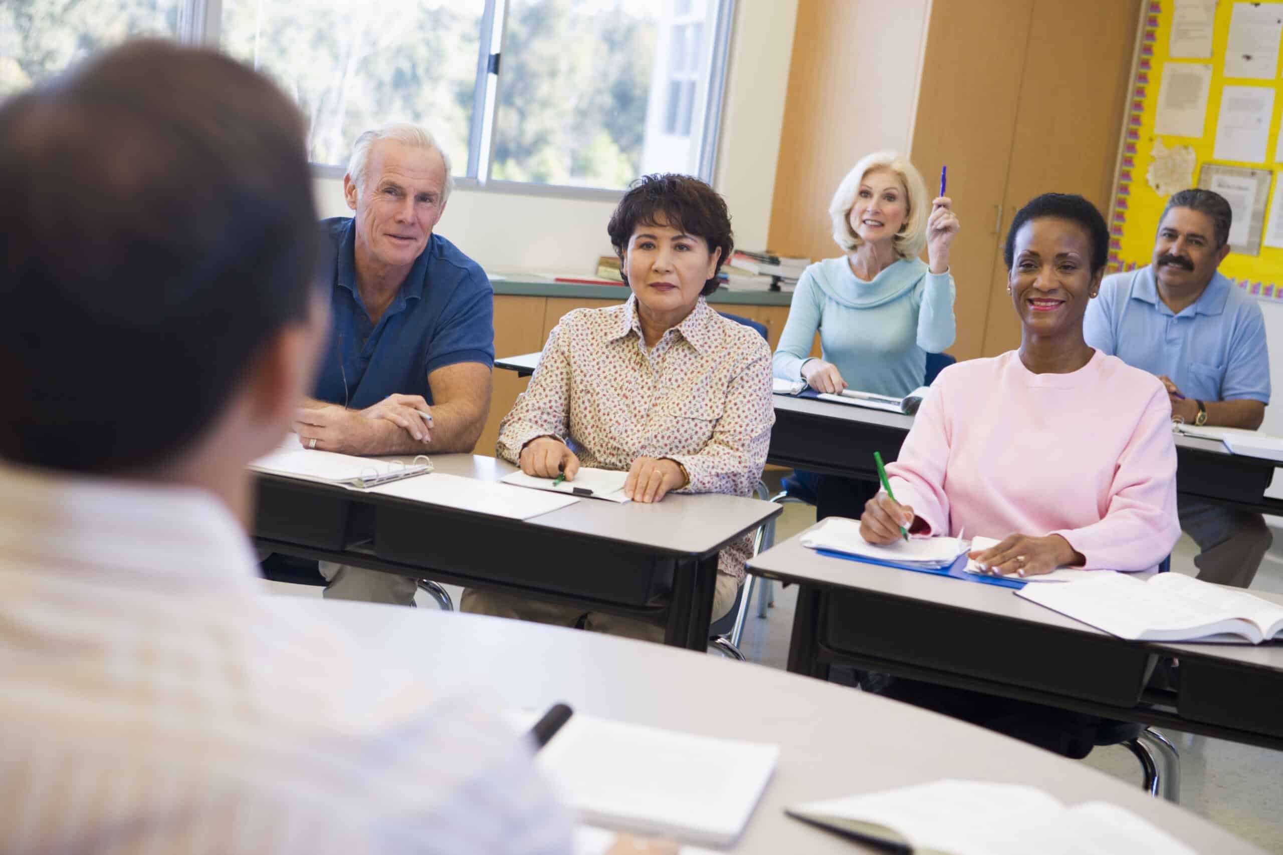Mature adults sitting at desks in classroom, woman raising hand to participate
