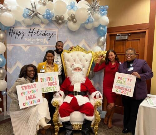 Kettering Health staff posing with Santa holding holiday-themed signs at festive balloon-decorated event