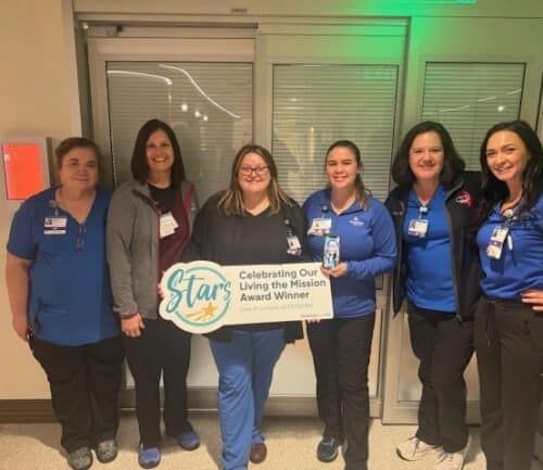 Six healthcare workers celebrating with "Living the Mission Award" banner in hospital hallway