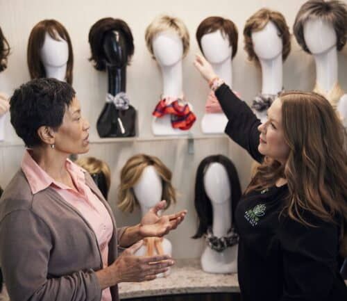 Two women selecting wigs from display wall of various wig styles on mannequin heads