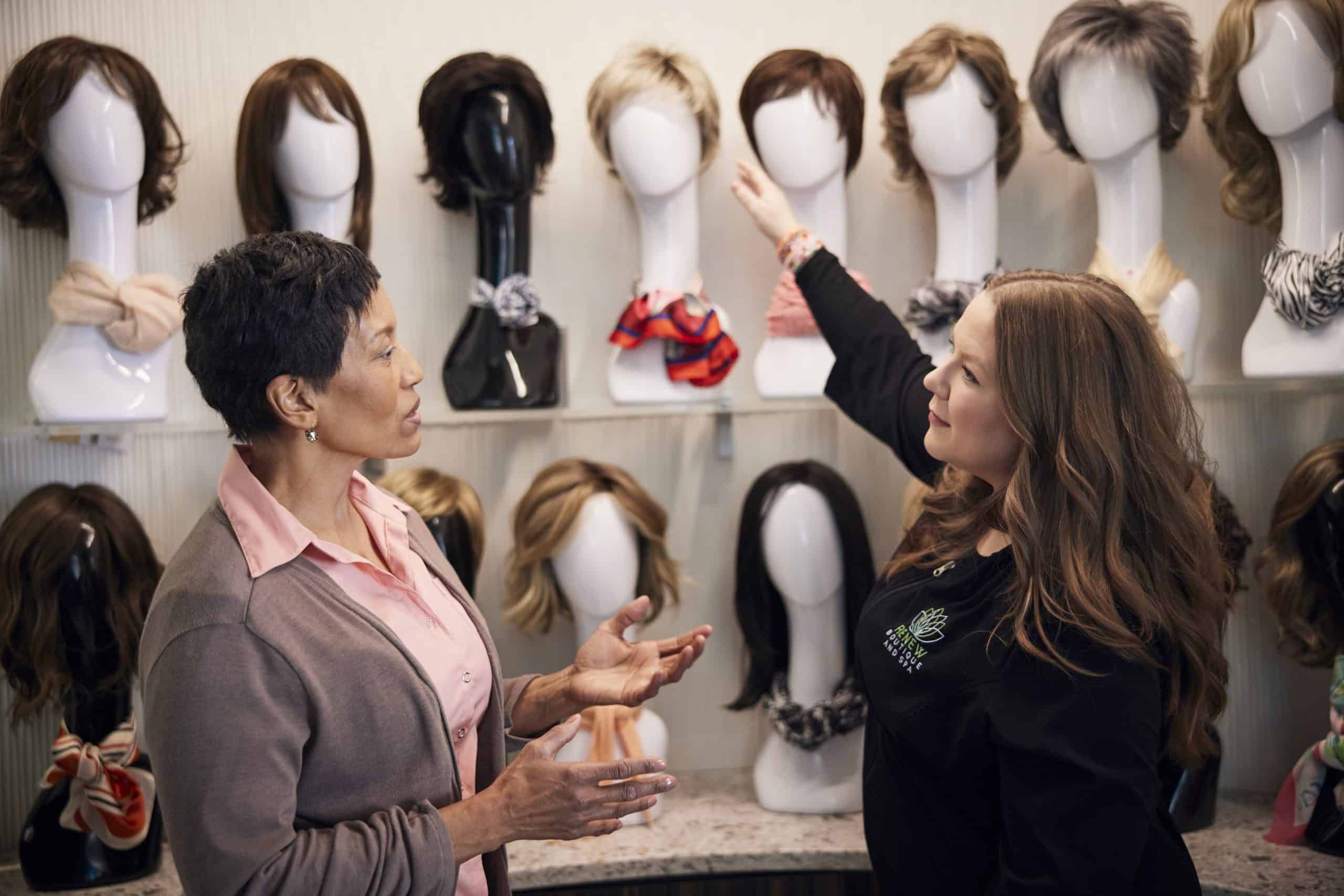 Two women selecting wigs from display wall of various wig styles on mannequin heads