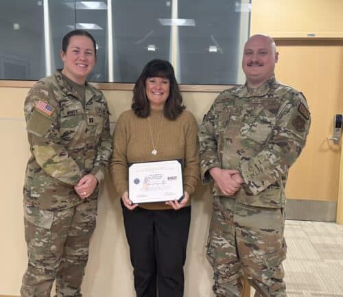 Three people posing with certificate, two in military uniforms flanking woman in civilian clothes
