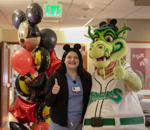 Healthcare worker poses with Disney mascot and dragon mascot holding balloons in hospital hallway