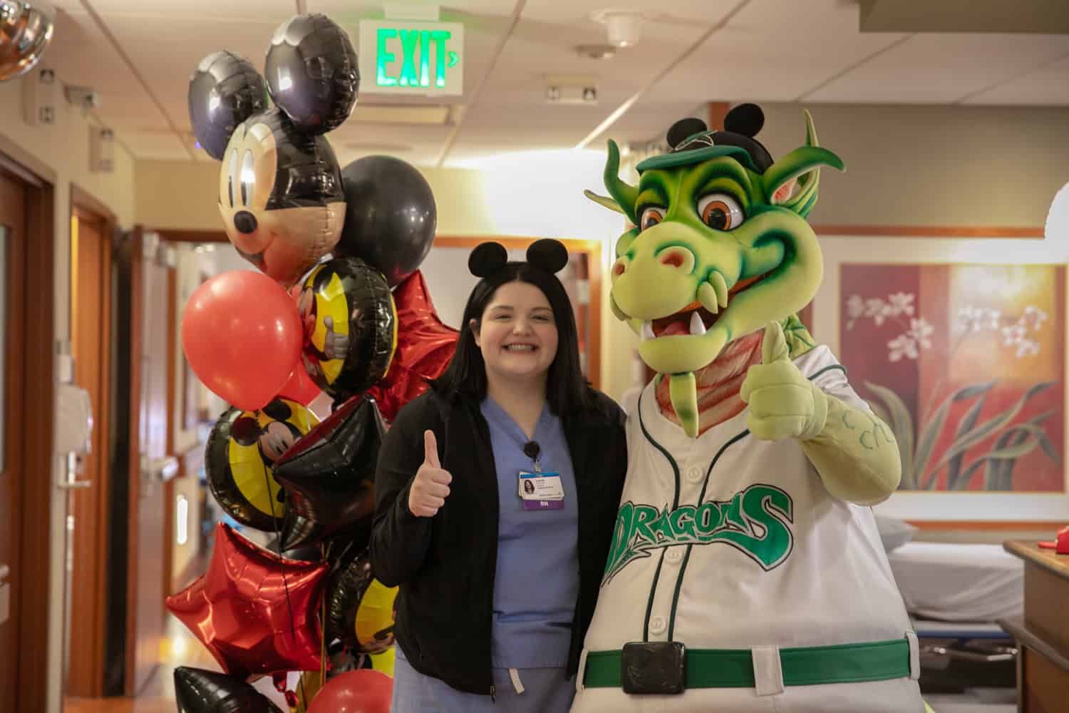 Healthcare worker poses with Disney mascot and dragon mascot holding balloons in hospital hallway