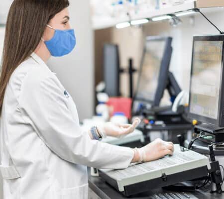 Female pharmacist wearing blue mask and white coat working at computer in clinical pharmacy setting.