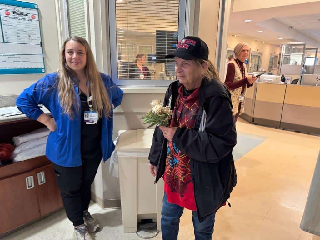 Staff member in blue jacket welcomes visitor holding flowers at Kettering Health facility reception area.