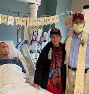 "Patient in hospital bed receives visit from two supporters wearing ""Congratulations"" banner in background."