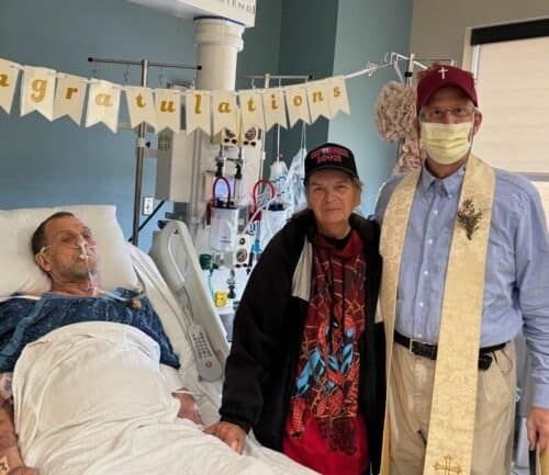 "Patient in hospital bed receives visit from two supporters wearing ""Congratulations"" banner in background."