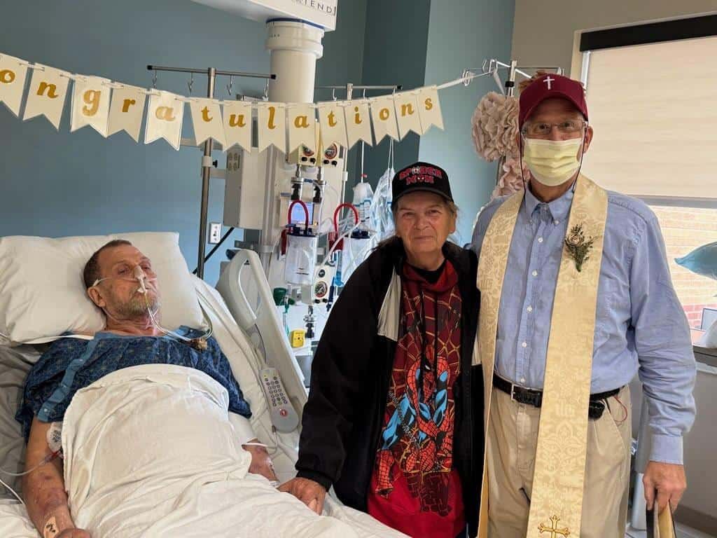 "Patient in hospital bed receives visit from two supporters wearing ""Congratulations"" banner in background."